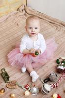 Little girl in white blouse and pink chiffon skirt is playing with Christmas decorations on a beige knitted plaid. photo