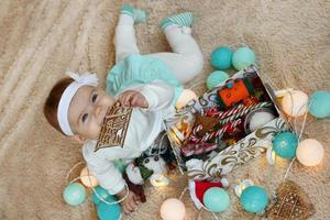 hermosa niñita con vestido azul y blanco está acostada sobre una manta beige, jugando con adornos navideños y mirando a la cámara. foto