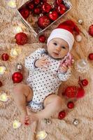 Adorable little girl in Santa Claus red hat is playing with wooden toy on a beige plaid with red and white Christmas decorations and Christmas lights, top view. photo