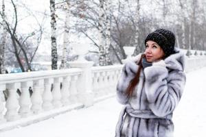 joven hermosa mujer con pelo rojo y ojos azules en abrigo de piel sintética gris con capucha está caminando en el parque nevado de invierno. foto