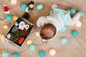 Little girl is playing with blue and white Christmas lights and box with Christmas decorations on a beige plaid, top view. Waiting for New Year and Christmas holidays. photo