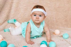 Cute little girl with beautiful blue eyes in blue dress is sitting on a beige plush plaid with Christmas lights. photo