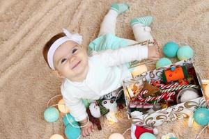 hermosa niñita sonriente con vestido azul y blanco está acostada sobre una manta beige, jugando con adornos navideños y mirando a la cámara. foto