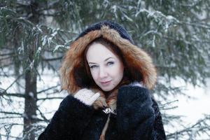 joven mujer hermosa sonriente con ojos azules en abrigo de piel sintética negra con capucha roja en el fondo del parque nevado de invierno. foto