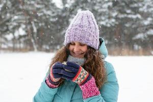 bella mujer sonriente con el pelo largo y rizado con chaqueta azul de invierno y guantes coloridos tejidos sostiene en sus manos una taza de té caliente en un paseo por el bosque nevado de invierno. foto