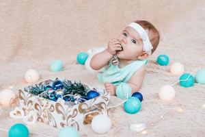 Cute little girl in blue and white dress is playing with Christmas decorations from a box on a beige plaid with Christmas lights. photo