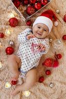 linda niña sonriente con sombrero rojo de santa claus está jugando con un juguete de madera en una manta beige con adornos navideños rojos y blancos y luces navideñas, vista superior.