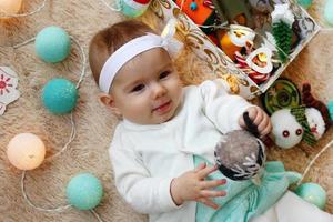 Beautiful smiling little girl in blue and white dress is playing with felt ball on a beige plaid with Christmas decorations and lights, top view. photo