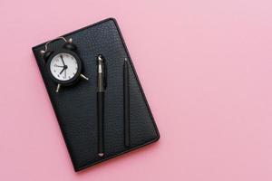 Black notepad with a pen and an alarm clock on a pink background. Using photo