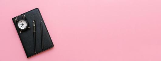 Black notepad with a pen and an alarm clock on a pink background. Using photo