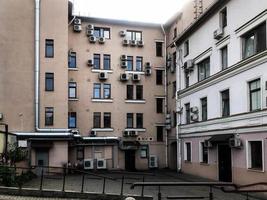 an orange house stands in the city, with many white air conditioners hanging from the windows. there is another house nearby. a pipe for draining water into the sewer hangs on the side photo