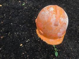 a bright orange construction helmet rests on the black fertile ground during construction. a new complex is under construction in the city center. green young plants grow next to the helmet photo