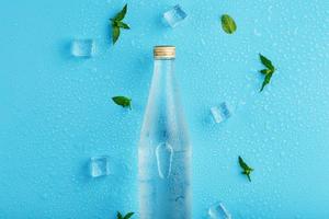botella de agua fría, cubitos de hielo, gotas y hojas de menta sobre un fondo azul. foto