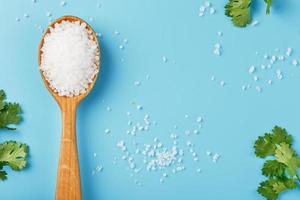 Sea salt in a wooden spoon on Large salt crystals with greens on a blue background. photo