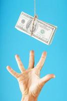 una mano intenta agarrar un fajo de dinero en una cuerda sobre un fondo azul. los billetes de un dólar cuelgan de una cuerda. foto