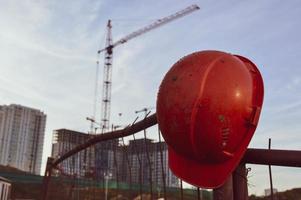 casco naranja brillante cuelga de la valla. en el fondo, una grúa de pórtico de construcción para mover objetos pesados a edificios altos. construcción de nuevo complejo residencial. pintura brillante en el casco foto