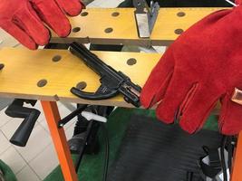 A welder man in red work gloves is working and holding an electron and clamps for welding at the table photo