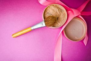 Beauty box, friable matte mineral powder with a special brown beautiful wooden brush from natural nap for a make-up on a pink violet background with a satin delicate ribbon. Flat lay. Top view photo