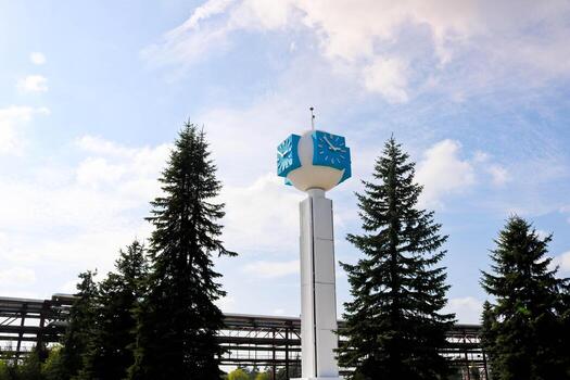 Unusual clock on a background of a white-blue sky. A clock in the form of a molecule, an atom. On chemical production, a watch-column, a clock tower in a spruce forest. Backdrop of chemical pipelines photo