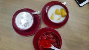 Top view of Thai dessert with strawberry in syrup, banana in coconut milk and taro Dumplings in coconut cream in red bowl and mobile phone or telephone on wooden table or background. Sweet Asian food. photo