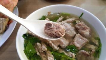 Hand holding spoon and scoop pork ribs or bone of Vietnamese Rice Noodle Soup with pork spare ribs and fresh vegetable on white bowl at Vietnam restaurant. Asian food and Famous in Vietnam. photo