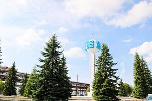 Unusual clock on a background of a white-blue sky. A clock in the form of a molecule, an atom. On chemical production, a watch-column, a clock tower in a spruce forest. Backdrop of chemical pipelines. photo