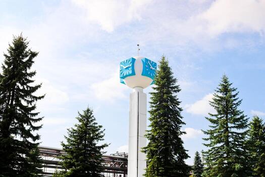 reloj inusual sobre un fondo de un cielo blanco-azul. un reloj en forma de molécula, un átomo. sobre la producción química, una columna de vigilancia, una torre de reloj en un bosque de abetos. telón de fondo de tuberías químicas. foto