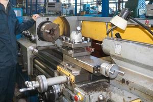 A man working in a robe, overalls stands next to an industrial lathe for cutting, turning knives from metals, wood and other materials, turning, making details and spare parts at the factory photo