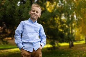 portrait of a cheerful boy in a blue shirt standing with his hands in his pockets against the backdrop of an autumn forest photo