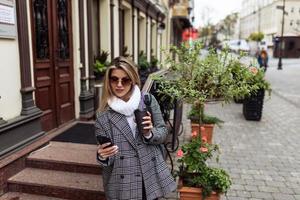 mujer viajera con estilo vestida de otoño con un vaso de café mira una ruta para dar un paseo en un teléfono móvil foto