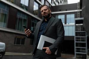 an adult male lawyer in a classic suit with a laptop in his hands and a mobile phone looks at the camera with surprise, Concept of a successful realtor photo