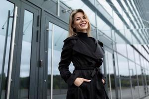 charming businesswoman in a black business coat at the door to the office building looking at the camera with a smile photo