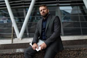 serious adult man with a laptop in his hands looks at the camera near the office building photo