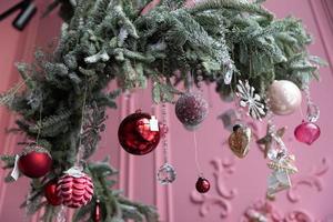 elegante arreglo de árbol de navidad de agujas de pino natural y abeto colgando del techo con adornos navideños contra el fondo de una pared rosa foto