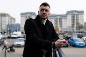 portrait of a brutal young businessman in a stylish black coat with a phone in his hands on the background of the city photo