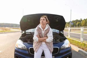 frustrated female driver sits by a broken down car on the hood waiting for help, transport auto insurance concept photo