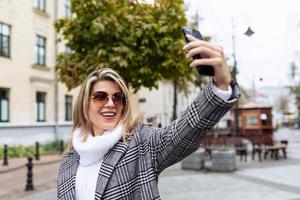 an adult woman in a white sweater and sunglasses makes a selfie on the phone on the background of the city photo