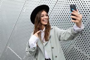 a young woman in a stylish coat and black hat speaks via video link against the background of an aluminum facade of a city building photo