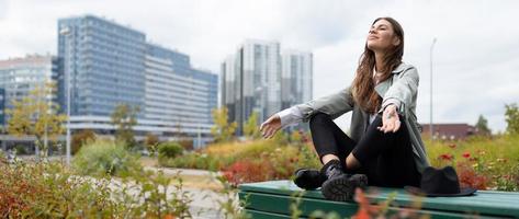 una joven durante una pausa para el almuerzo se relaja sentada en una posición de yoga en el parque al fondo de los edificios de oficinas foto