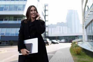 stylish businesswoman with a laptop in her hands with a smile on her face speaks on a mobile phone on the background of the office center photo