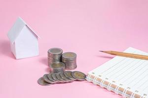 Top view image of notebook with pencil, stack of coins, house model on pink background photo