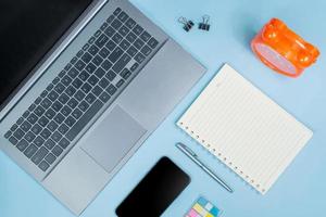 Top view of Office Table top with laptop computer, orange clock, notebook, smart phone, pen on blue table photo