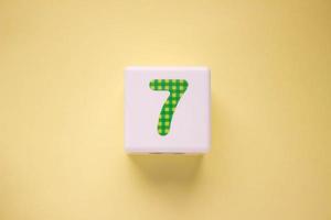 Close-up photo of a white plastic cube with a green number 7 on a yellow background. Object in the center of the photo