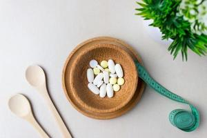 A handful of pills in a wooden plate with measuring tape. Surrounded by wooden spoons and green plant. The concept of weight loss, diet, medicine for weight loss. Top view photo