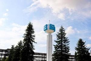 Unusual clock on a background of a white-blue sky. A clock in the form of a molecule, an atom. On chemical production, a watch-column, a clock tower in a spruce forest. Backdrop of chemical pipelines. photo