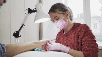 chica en los técnicos de uñas rosas una máscara médica y guantes haciendo manicura a un cliente en el salón de belleza. el cuidado de las uñas. video