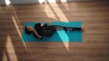 Athletic woman in sports overalls doing yoga and stretching while lying on a fitness mat at home on a wooden floor in bright sunlight. Top view. Slow motion. video