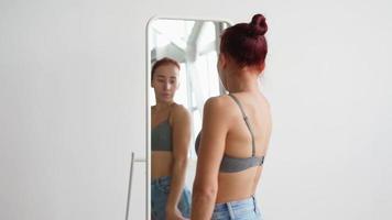 femme mince et heureuse en débardeur et jeans court jusqu'à miroir se regarder. jolie fille regarde dans le miroir en choisissant ce qu'elle porte à la maison sur fond blanc. sourit et s'amuse en profitant du bonjour. video