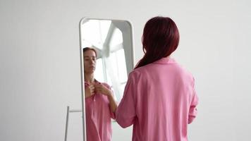 une jeune femme en chemise rose redresse ses cheveux roux près du miroir, se prépare pour le travail le matin. video