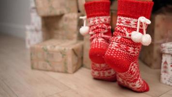 avvicinamento, bellissimo femmina gambe nel rosso nuovo anni calzini siamo adatto per decorazione un' Natale albero su in punta di piedi. nuovo anno, inverno atmosfera, caldo luce. festivo umore. video